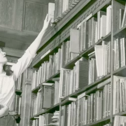 A young woman places a book at the very top of a bookshelf in a library. Black and white.