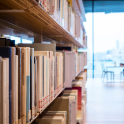 Close up of library books on a shelf, with a study table visible at the end of the aisle.